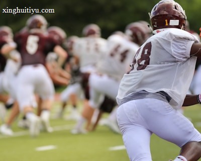 A group of football players wearing maroon and white uniforms are on a field. One player in the foreground, wearing a white jersey and red gloves, is in focus, while more players are visible in the background running during a game or practice.