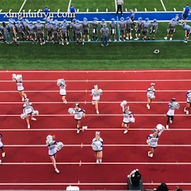 A group of cheerleaders is lined up on a red athletic track, performing routines with pom-poms. Behind them, a football team stands on a green field, dressed in gray and blue jerseys and helmets, observing the scene. Coaches and other officials are stationed along the sidelines.
