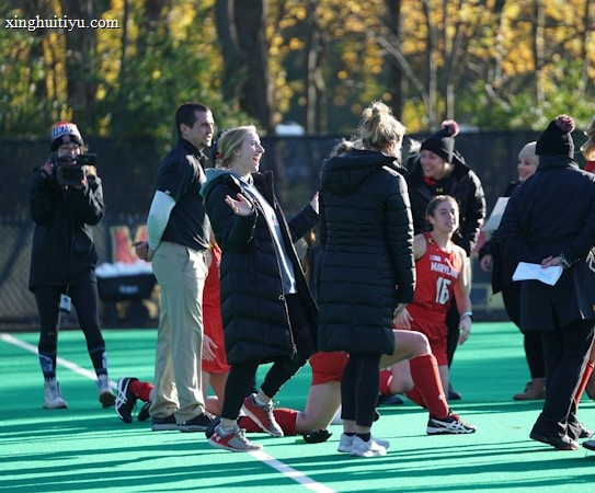 A group of people on a sports field, including several athletes in red uniforms and coaches wearing black coats. Some individuals are kneeling, while others are standing, engaged in animated conversation. A photographer or videographer is capturing the scene beside them. The background features a fence and autumn trees with yellow leaves.