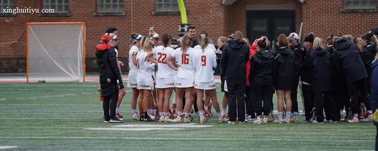 A group of athletes wearing sports uniforms huddles on a grassy field, likely preparing or strategizing for a game. The players are surrounded by additional team members or coaches wearing black jackets. A goalpost and net are visible in the background, along with a brick building.