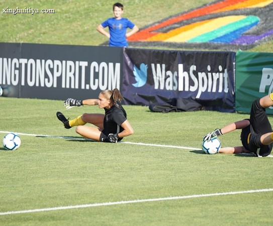 Two football players are performing a training drill on a grassy field. One is leaning back while controlling a soccer ball with her hands, and the other is lying on her back with her legs and arms up, holding another soccer ball. Both are wearing black attire with yellow socks. In the background, banners display social media handles and web addresses, and a person in a blue shirt stands nearby, watching the scene.