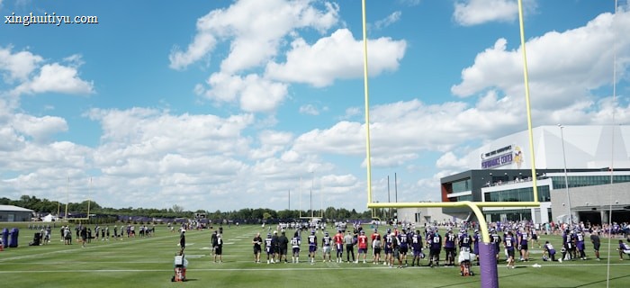 A group of football players, wearing uniforms, are lined up on a green field with a bright blue sky and white clouds in the background. There is a large sports facility building to the right, and the scene is framed by yellow goalposts. Equipment and a water cooler can be seen on the field.
