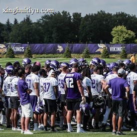 A group of football players huddled together on a grassy field, wearing jerseys and helmets. The players appear to be in a team meeting or discussion. Trees and a fencing banner with a team's name are visible in the background under a partly cloudy sky.