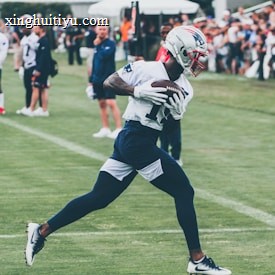 A football player wearing a helmet and uniform is running on a grass field while holding a football tightly. Surrounding him are other players and coaches, with a crowd of spectators in the background. The scene appears to be a practice or training session with people observing the action.
