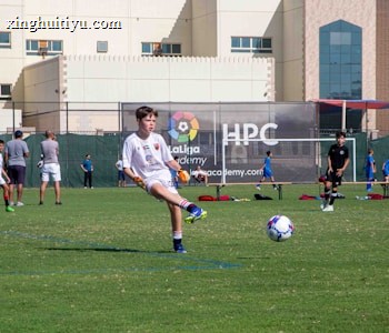 A youth soccer player in a white uniform prepares to make a pass during a practice or game on a green soccer field. Other players and coaches are present in the background, along with a large building and a banner with logos. There is sunlight suggesting a bright, clear day.