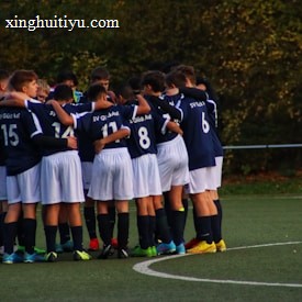 A group of young football players wearing navy blue jerseys and white shorts are huddled together on a green sports field. The players have their arms around each other, suggesting camaraderie and teamwork. The background consists of trees with autumn foliage, indicating an outdoor setting.