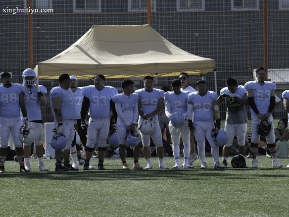 A group of football players in uniform standing in a line on a football field. They are wearing light blue jerseys, and some have helmets in their hands. A tent and goal nets are visible in the background.