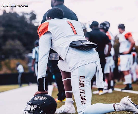 A football player wearing a white uniform with orange accents is kneeling on the grass next to a sideline, holding a black helmet with 'CAU' lettering. Other players and coaches are visible in the background.